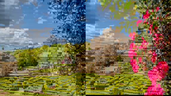 A large, grand house with a manicured hedge maze in the foreground, with blooming pink flowers framing the right side.