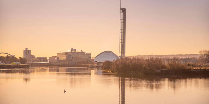A calm river at sunset with swans swimming, modern buildings, and a tall tower in the background.