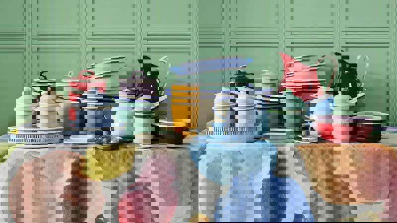 A collection of pastel and colourful enamel kitchenware, including a pitcher, cups, and bowls on a patterned tablecloth.