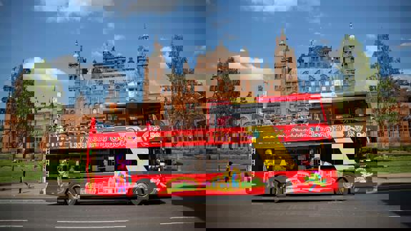 A bright red double-decker city sightseeing bus in Glasgow, with passengers on the top deck, passing the grand Kelvingrove Art Gallery and Museum.