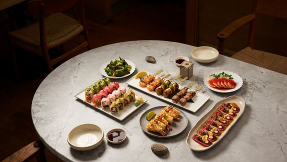 A round marble table set with assorted sushi rolls, skewered grilled items, sashimi, and small plates in a cosy restaurant.