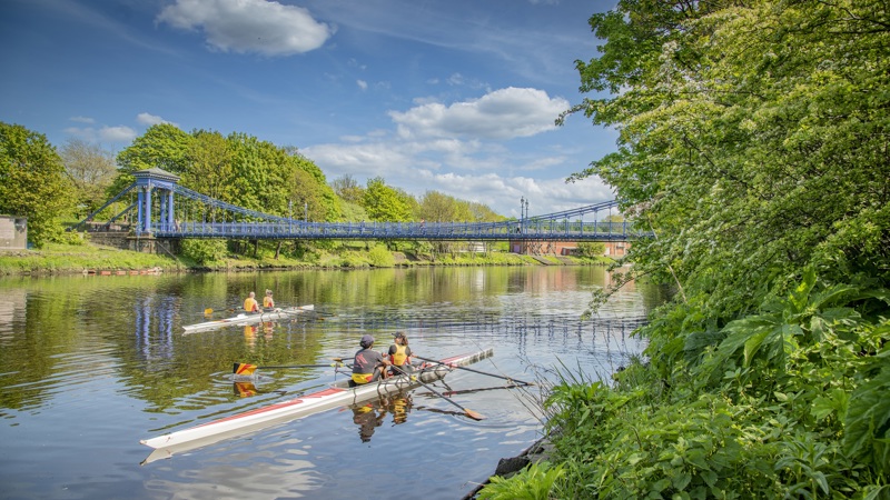 A canoeist rides along a still river on a sunny day and is about to go beneath an ornate blue and white bridge. Trees overhang the banks of the river.