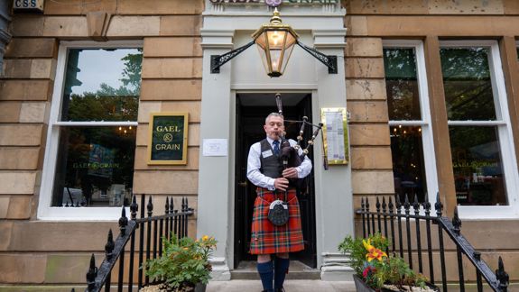 A person in a red tartan kilt plays bagpipes outside a stone building named "GAEL & GRAIN," which has a sign reading "CLOSED PRIVATE EVENT" on the door.