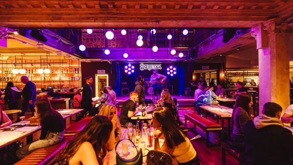 A lively bar scene with people seated at wooden tables under warm yellow lighting. A guitarist performs on stage with purple spotlights behind.