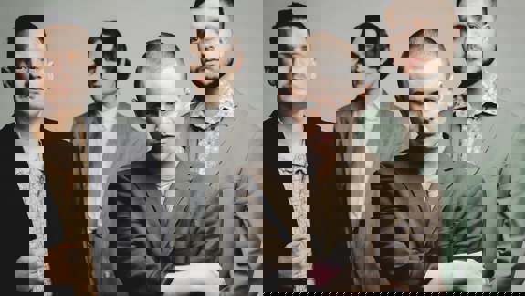 A close-up of four men in suits posing for a photo against a grey background.