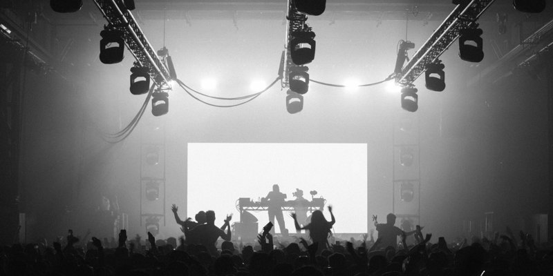 Silhouetted crowd at a concert cheering in front of a brightly lit stage with a DJ performing under overhead lighting rigs.