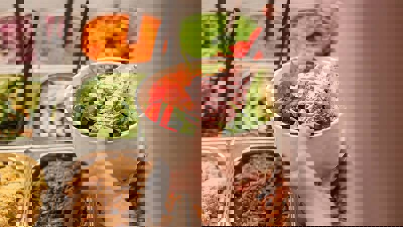 A person holds a white bowl filled with a fresh salad, including avocado and a pink sauce, over a buffet of colourful ingredients.