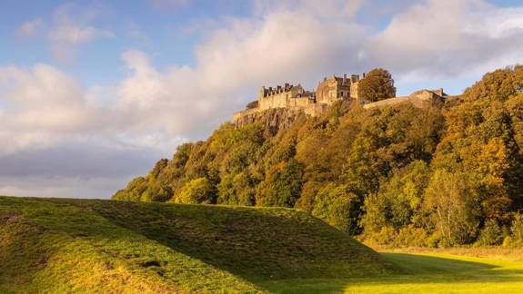 Stirling Castle sits majestically on a hilltop, surrounded by golden autumn foliage and a grassy mound in the foreground.