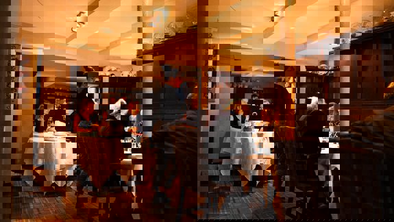 A warm, traditional restaurant interior with patrons dining at white-clothed tables and a server standing on a tartan carpet.