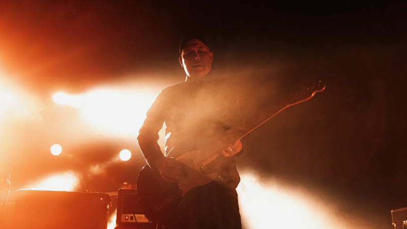 Stuart Braithwaite A musician silhouetted by bright orange stage lights plays an electric guitar amidst smoke or haze.