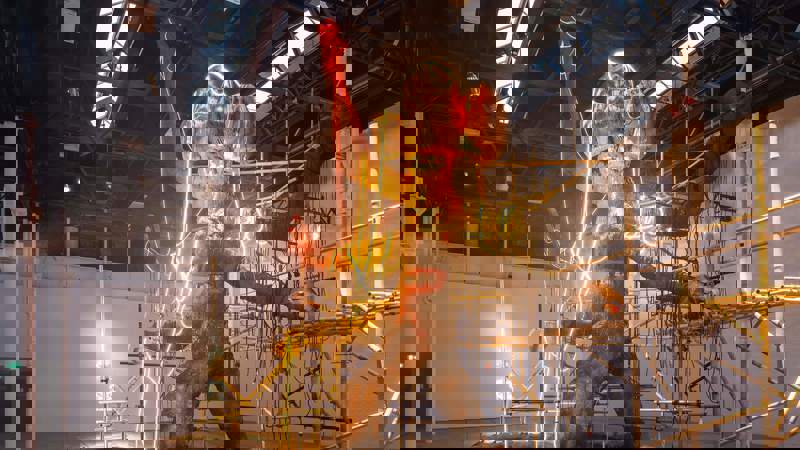 A huge, mud-like figure with an elephant's head stands in a shallow pool of water, illuminated by red and yellow neon lights, surrounded by scaffolding and sandbags.