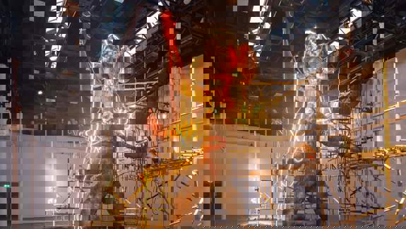 A huge, mud-like figure with an elephant's head stands in a shallow pool of water, illuminated by red and yellow neon lights, surrounded by scaffolding and sandbags.