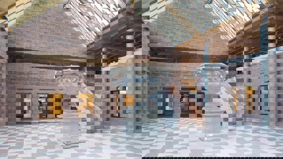 Interior view of a gallery space with red sandstone walls, a checkered floor, and a large decorative stone urn displayed in the centre beneath a sloped glass roof.