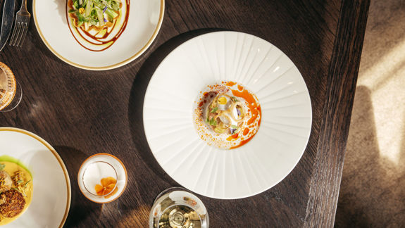 Overhead shot of small, elegantly plated dishes on a dark wooden restaurant table, with wine glasses.