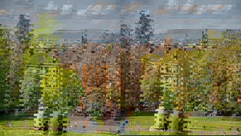 Two people sit on a grassy hill, looking out at a city skyline with buildings and distant hills under a cloudy sky.