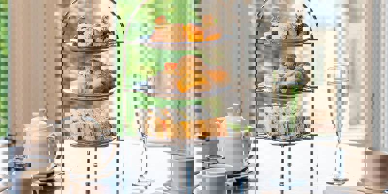 A three-tiered stand featuring finger sandwiches, scones, and small cakes, set on a dark wood table with a teapot and a glass of champagne against a window view.