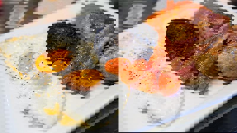 A full English breakfast features two fried eggs with runny yolks, roasted cherry tomatoes, a large Portobello mushroom, bacon, and sausages, all served on a white rectangular plate.