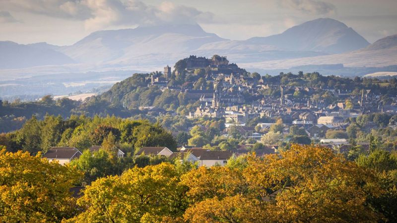  A view of Stirling Castle perched on a hill above the city, with a backdrop of rolling hills under a cloudy sky. Autumn trees are in the foreground.