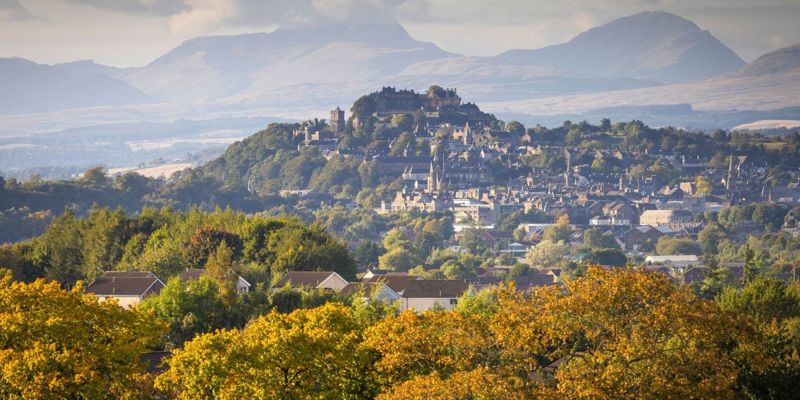 A view of Stirling Castle perched on a hill above the city, with a backdrop of rolling hills under a cloudy sky. Autumn trees are in the foreground.
