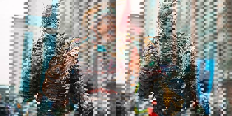 A person in an elaborate, fantastical costume and headdress made from plastic bags, bicycle tires and bits of trash, is performing outdoors, with tall, urban buildings in the background.