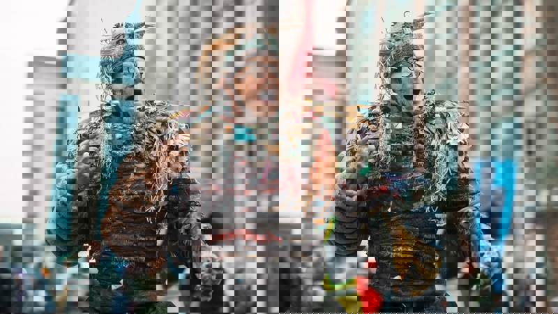 A person in an elaborate, fantastical costume and headdress made from plastic bags, bicycle tires and bits of trash, is performing outdoors, with tall, urban buildings in the background.