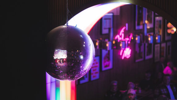 Hanging disco ball reflecting colourful lights in a night-time bar interior with illuminated arch, pride flag, and framed wall art.