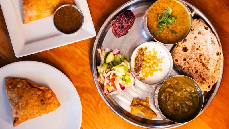Overhead shot of a metal platter with Indian dishes, including curries, salad, roti, and samosas with chutney. Colourful, detailed presentation of traditional cuisine.