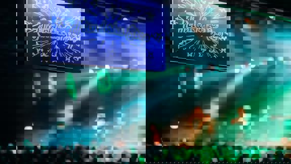 A close-up of a blue and white neon sign inside the venue that reads "Mind the Step" surrounded by the signature Barrowland starburst design, with a blurred concert crowd and stage lights below.