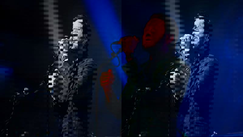 A male singer passionately performing on stage with a microphone under dramatic stage lighting.