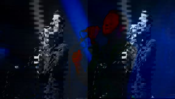 A male singer passionately performing on stage with a microphone under dramatic stage lighting.