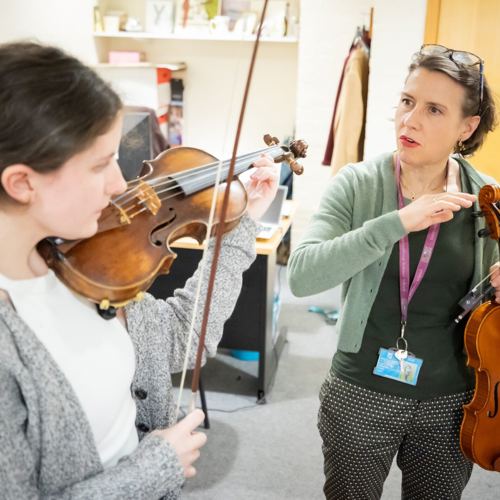 A young violinist plays attentively while receiving instruction from a teacher holding a violin. The setting is a cosy music room, creating a focused atmosphere.