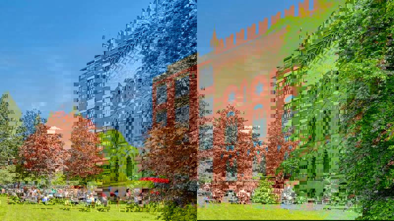 A large red brick building, West Brewery, with a beer garden on the grass in front of it filled with people.