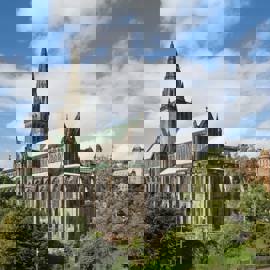 The grand exterior of Glasgow Cathedral with its spire reaching into a blue sky with white clouds, surrounded by green trees.