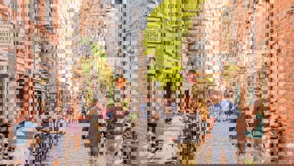 A bustling cobbled lane with outdoor dining, string lights, and historic brick buildings under a bright sky.