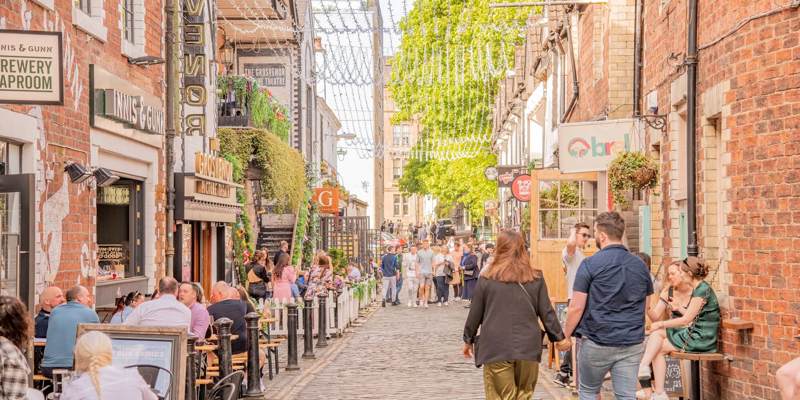 A bustling cobbled lane with outdoor dining, string lights, and historic brick buildings under a bright sky.