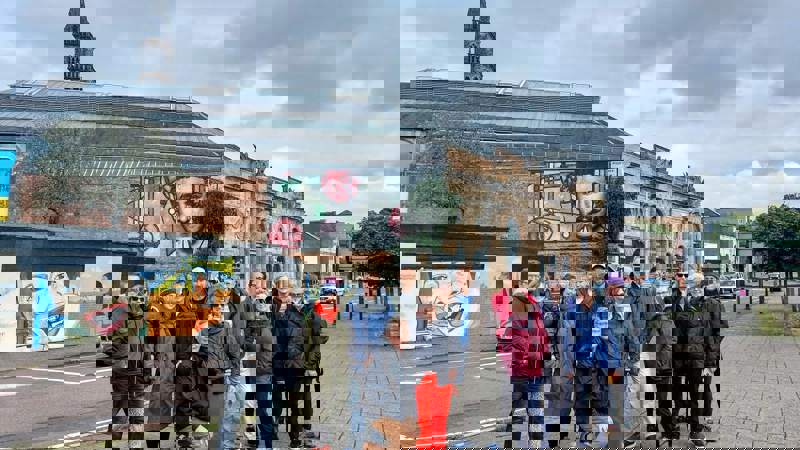 A large group of people smiling on a sidewalk in front of a brick building with a large mural and a steeple.