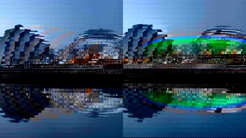 The SSE Hydro and SEC Armadillo are reflected in the water at night. The SSE Hydro is lit up with green and blue lights.