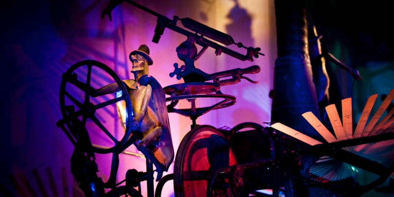 Artistic mechanical sculpture with metallic figures and gears illuminated by colourful lights in a dramatic, surreal setting.