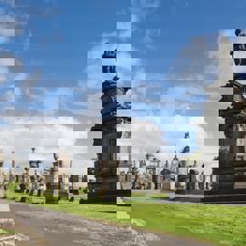 A wide shot of the Glasgow Necropolis, a Victorian cemetery with many ornate stone monuments, set on a green hillside under a blue, cloudy sky.