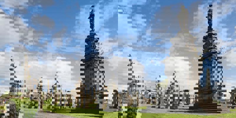 A wide shot of the Glasgow Necropolis, a Victorian cemetery with many ornate stone monuments, set on a green hillside under a blue, cloudy sky.