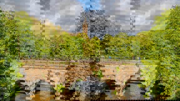 A stone bridge spanning a river, framed by dense greenery, with the spire of the University of Glasgow visible above the trees against a partly cloudy sky.