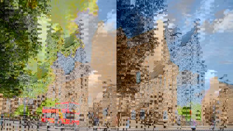 A large, stone building with a castle-like appearance stands on a street corner with a red double-decker bus passing by.