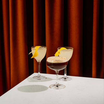 Three elegant cocktails on a white table against a backdrop of rich, red velvet curtains and a plush red chair.
