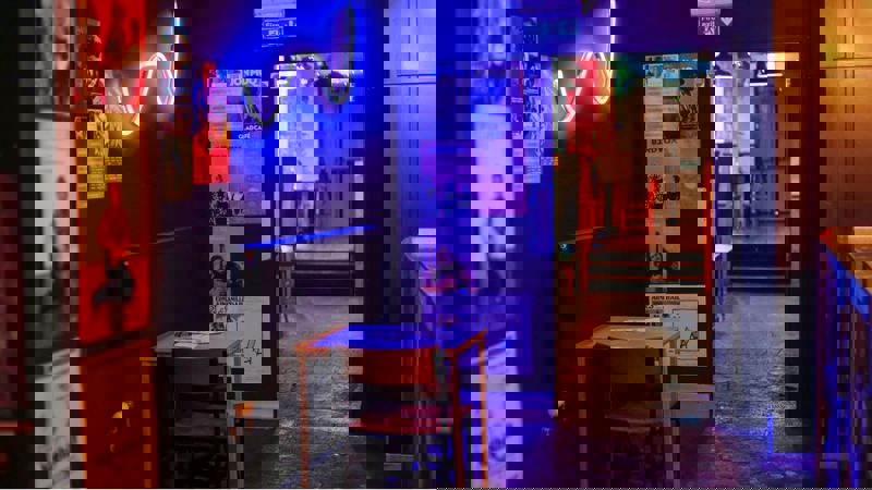 A small cafe with a dark interior, featuring a neon light sign on the wall and a small table and chair in the foreground.