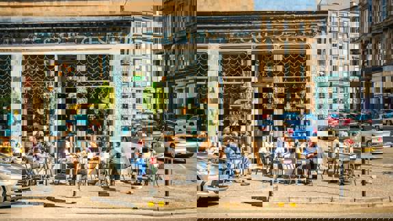 People sit at outdoor tables on a sunny day in front of a cafe with large windows and hanging plants.