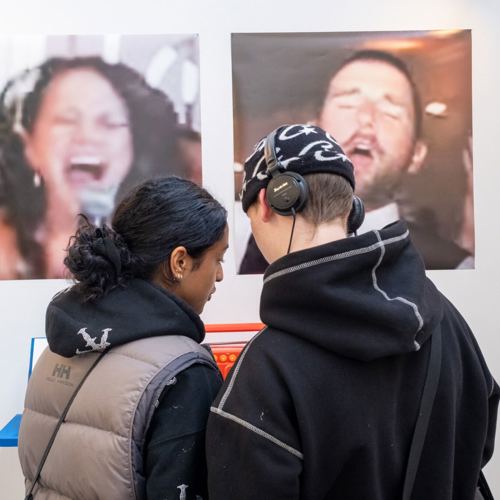 Two people stand closely, engaging with an exhibit. One wears headphones, the other looks on. Behind them are two blurred posters, showcasing singers mid-performance.