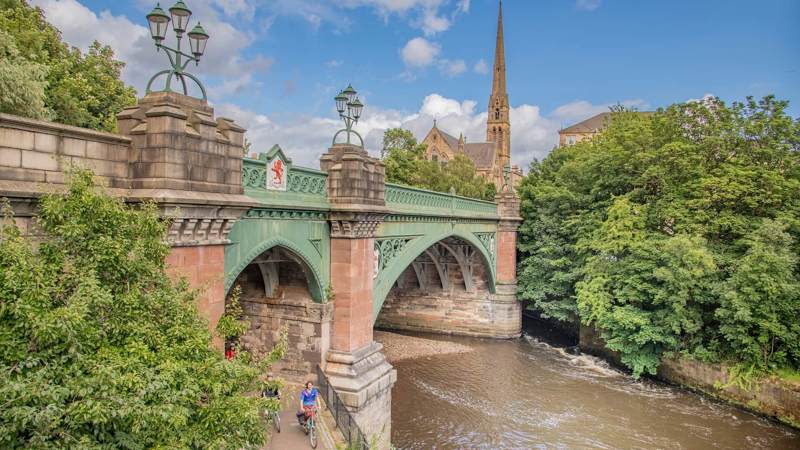 Historic stone and iron bridge over the River Kelvin in Glasgow, surrounded by lush greenery, with cyclists on the riverside path and a church spire in the background.