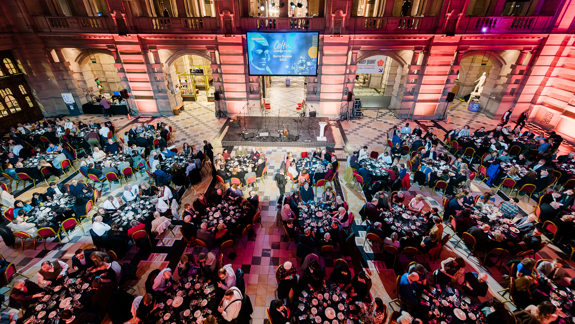 Large formal dinner event in a grand hall with round tables, guests seated, and a stage with a big screen at the front.