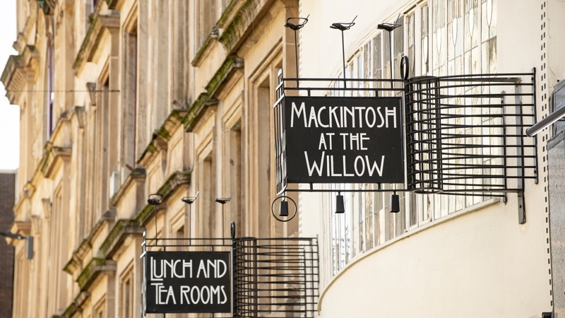 The exterior of 'Mackintosh at the Willow' on a building with a 'Lunch and Tea Rooms' sign and a 'Mackintosh at the Willow' sign.