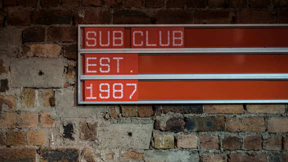  A club's changeable sign with white lettering reading "SUB CLUB EST. 1987" in three rows is mounted on a rough brick and concrete wall.